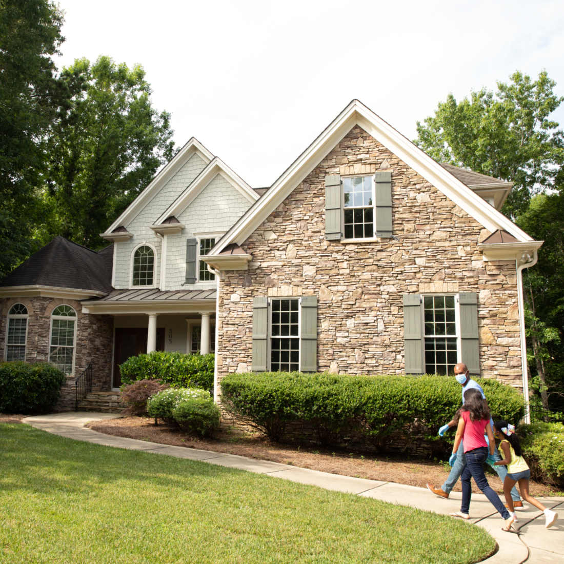 Parents [male, female] and child [female] walk up sidewalk towards entrance of large brick colonial style home