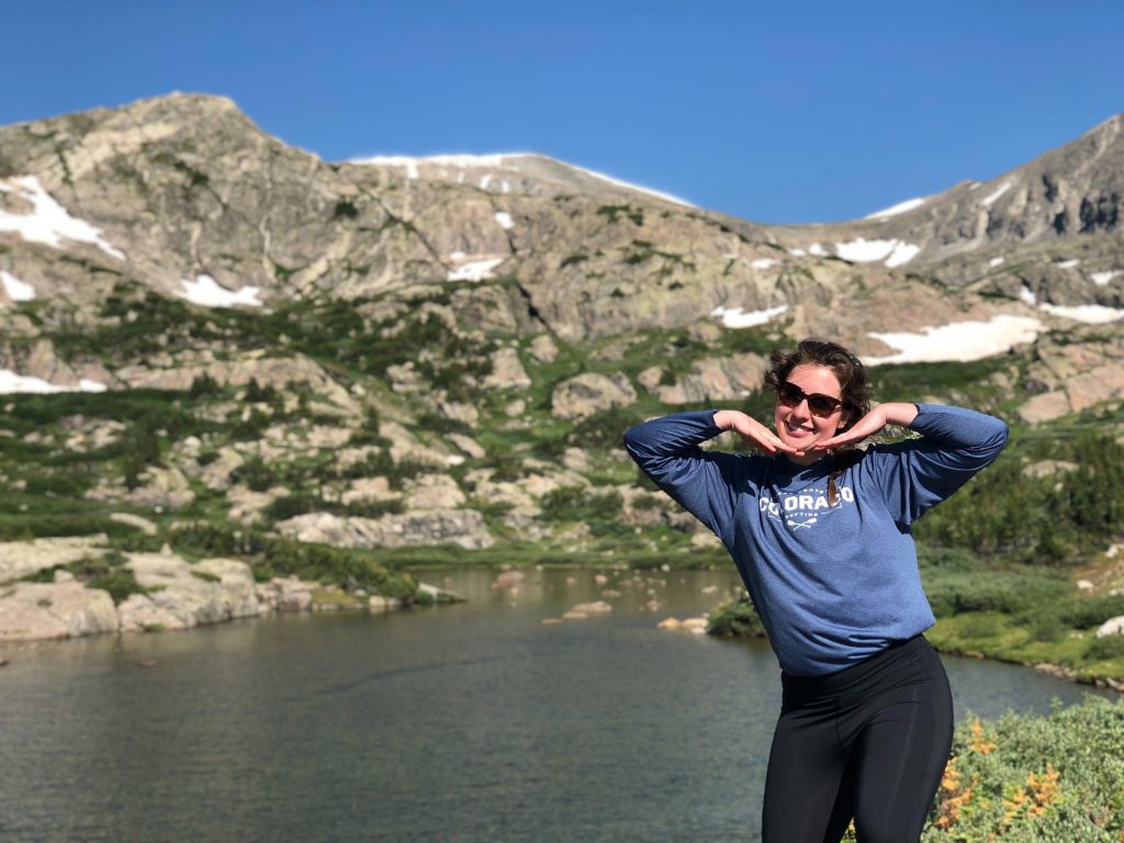 Woman standing in front of lake with mountain range in the background. She holds her hands below her chin to make the shape of a smile.