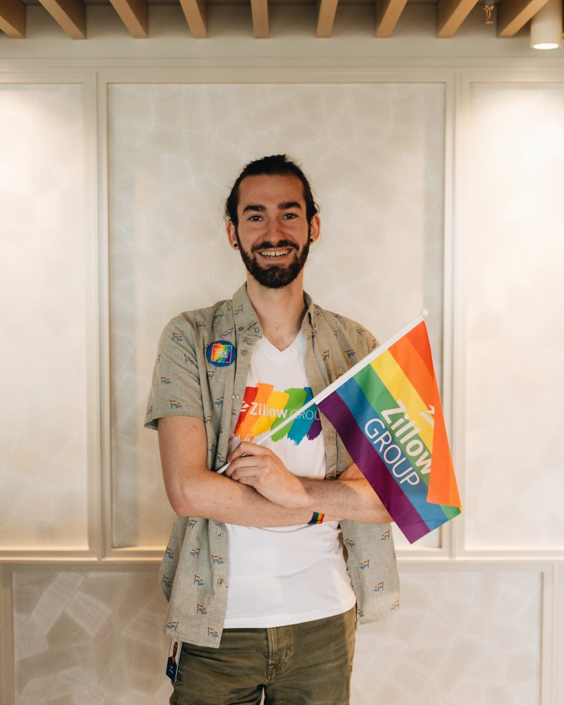 Portrait of software developer Jake Piccini, wearing a Zillow Pride network t-shirt and holding a small Pride flag