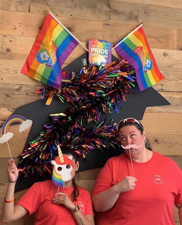 Snapshot of two people wearing masks with PRIDE flags hung on the wall behind them.