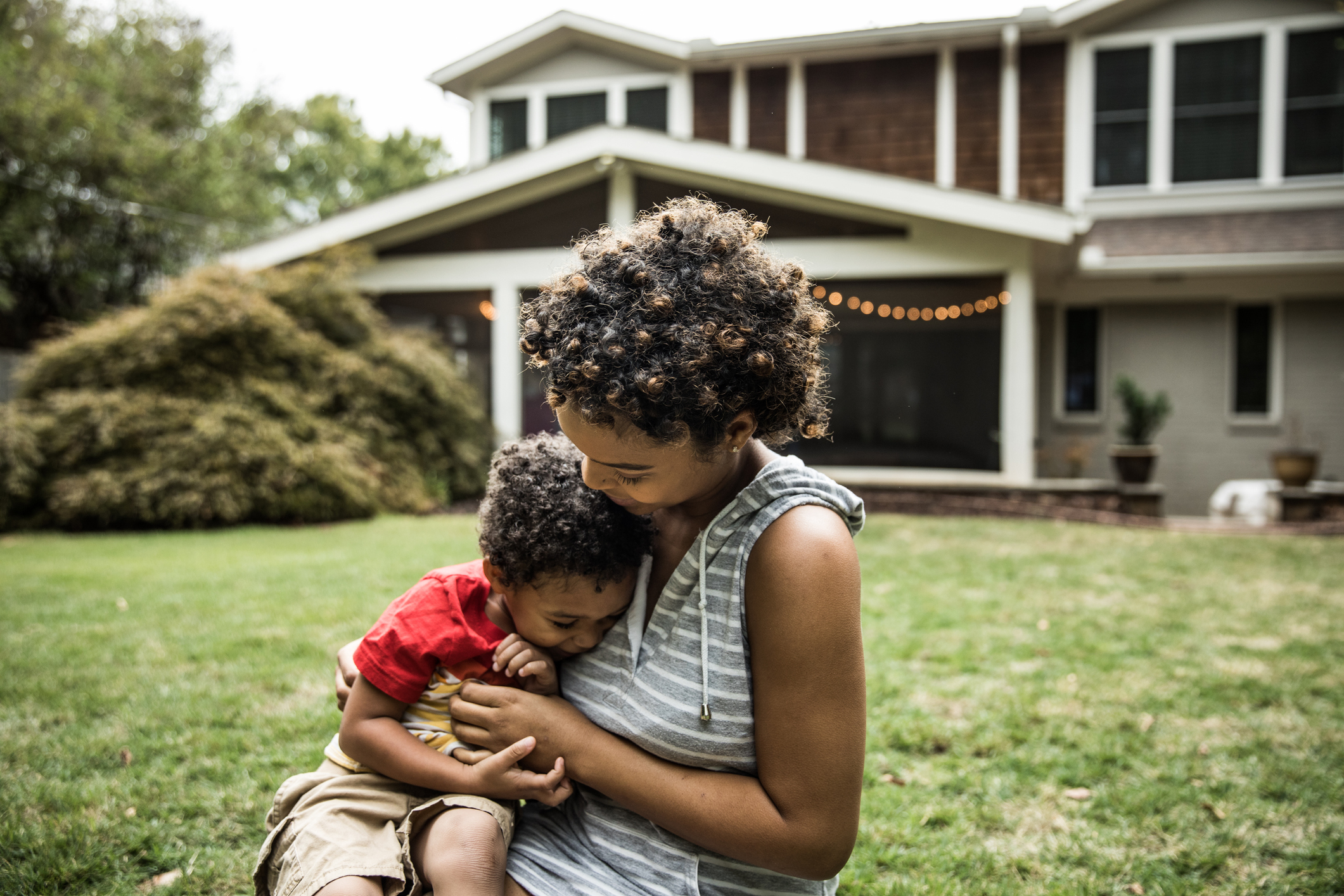 mother, toddler sit in grass in front of 2-story house