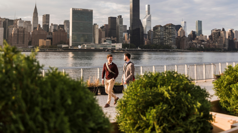 Two men walking along East River with NYC skyline in background