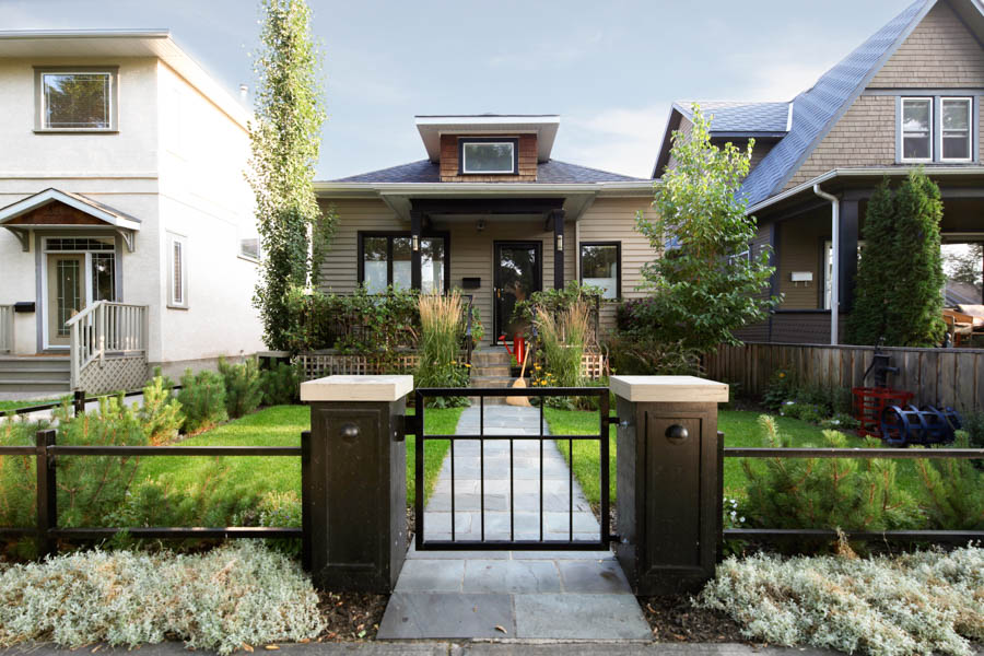 A simple but charming little house flanked by two taller houses. The house has a green lawn, a porch and a low iron fence.