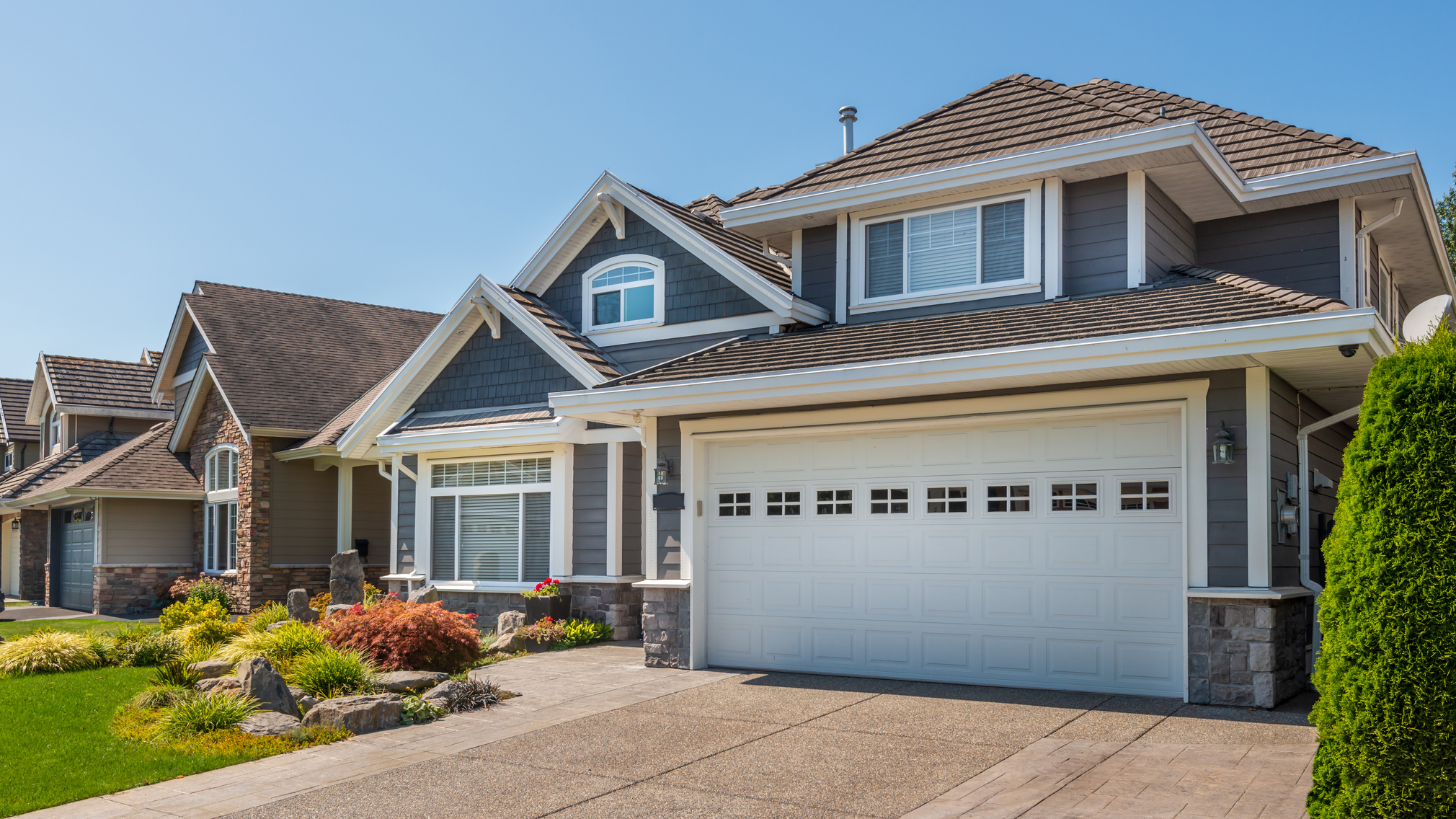 A dark gray, two-story house with white garage and plants outfront.