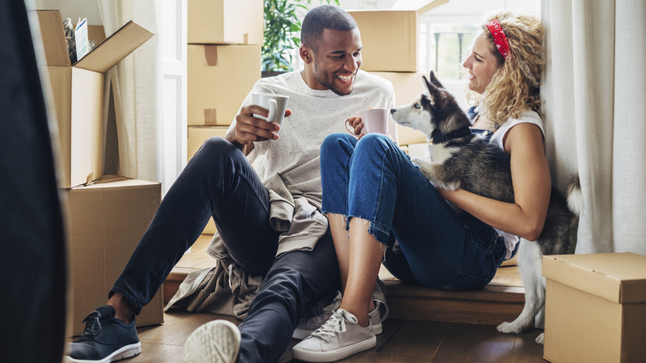 Man and woman drinking out of mugs with their husky puppy, surrounded by moving boxes.
