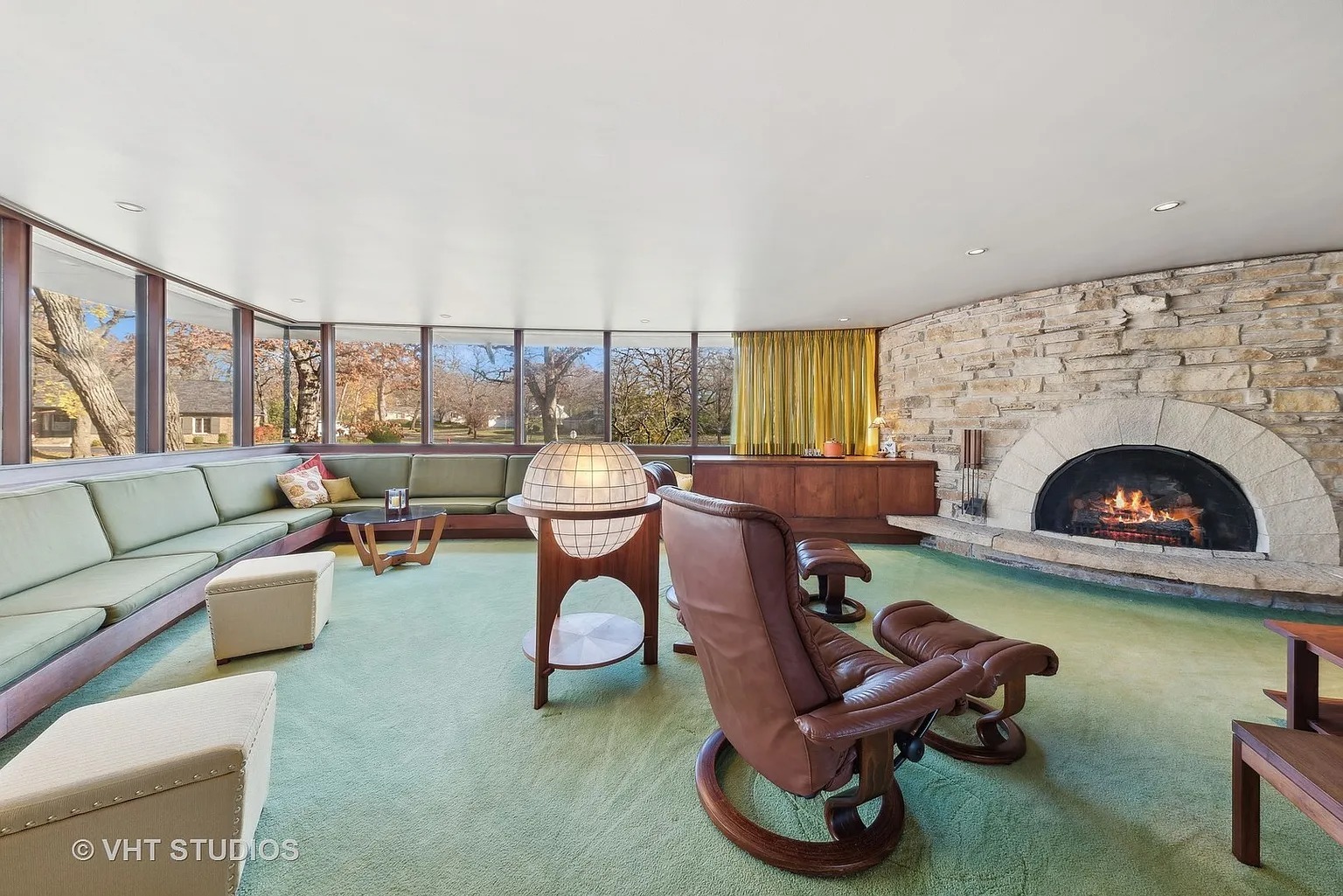 Step into the retro-futuristic living room, where curved lines, a globe lamp, and an olive green color palette create a midcentury modern vibe. The stone fireplace and large windows looking out to the lush greenery add a touch of warmth and natural light.
