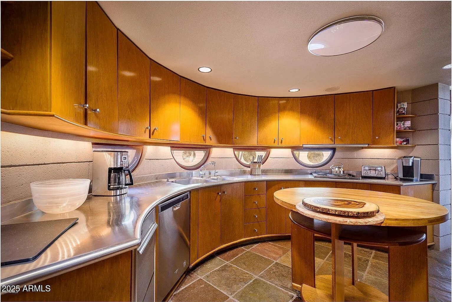 This kitchen is a throwback to the future, with its curved wood cabinetry, porthole windows, and a smooth metal countertop. The warm, earthy floor tiling is a surprising yet complementary touch to the space-age design.