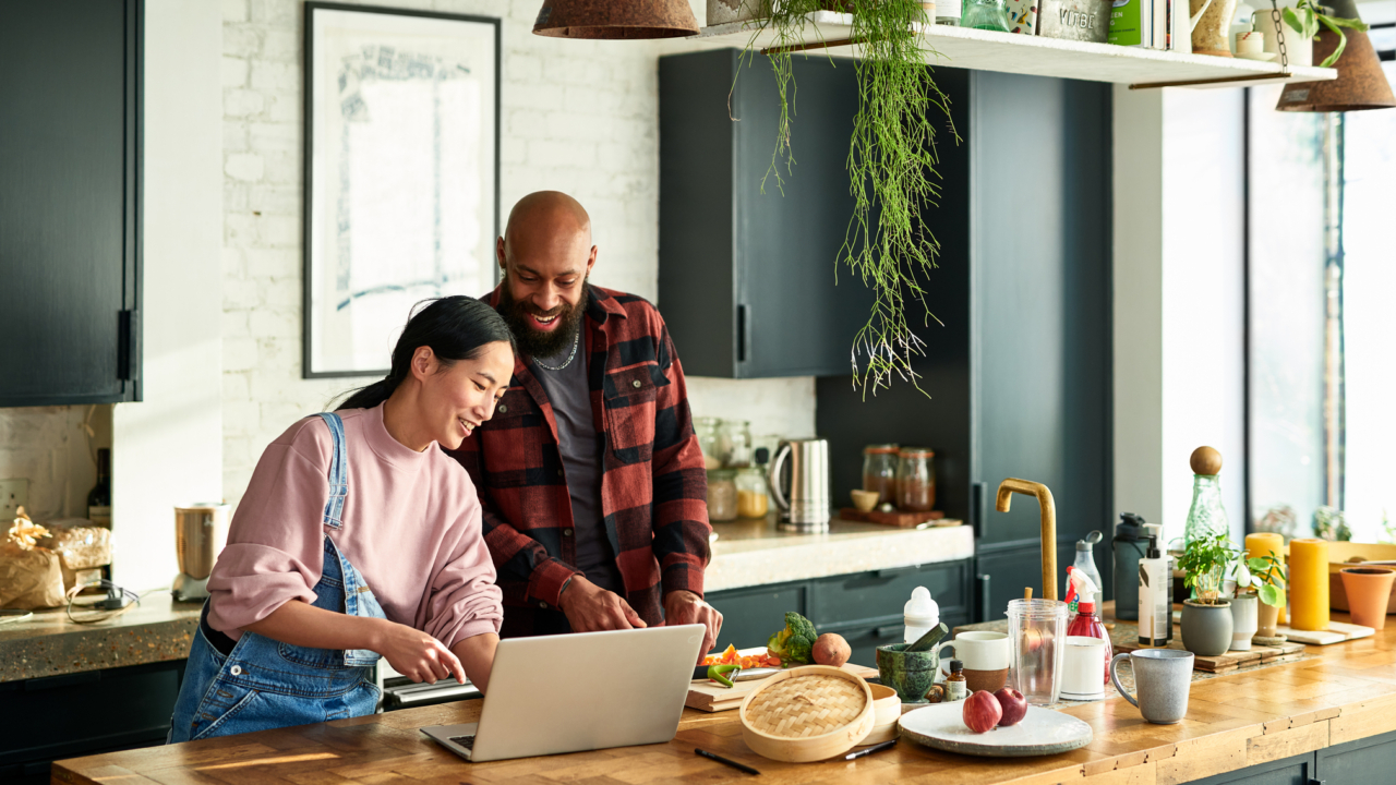 Smiling couple preparing food and using laptop to research how to shop for a mortgage without hurting their credit.