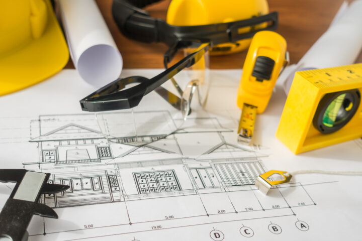 Image shows yellow construction helmet and drawing tools on blueprints.