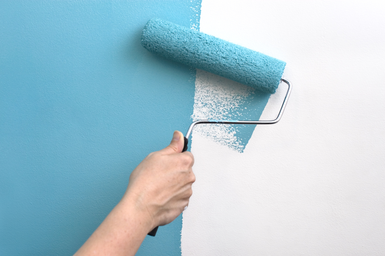 A hand with a paint roller covered in teal blue paint, painting over a white wall.