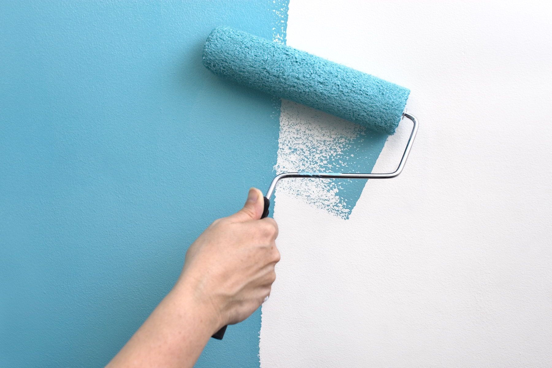 A hand with a paint roller covered in teal blue paint, painting over a white wall.