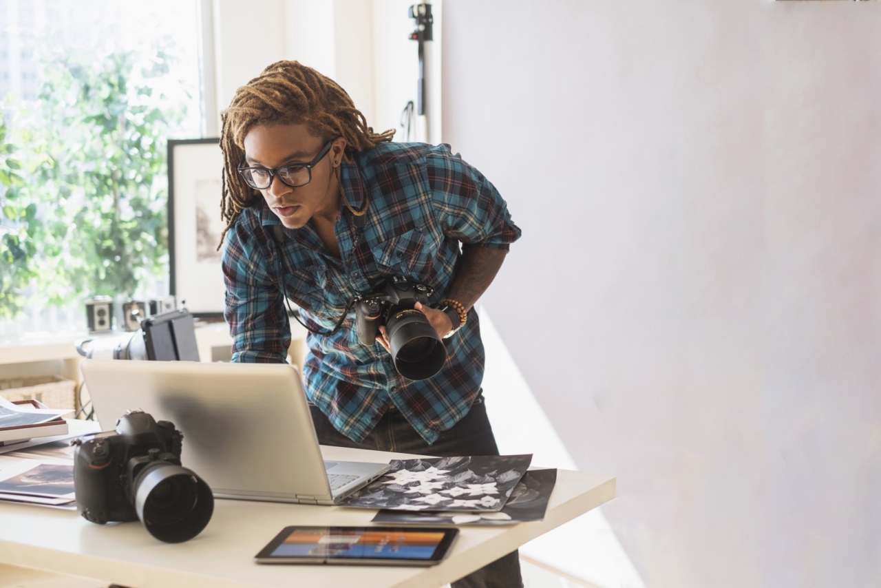 A photographer holds a camera while looking at his laptop.