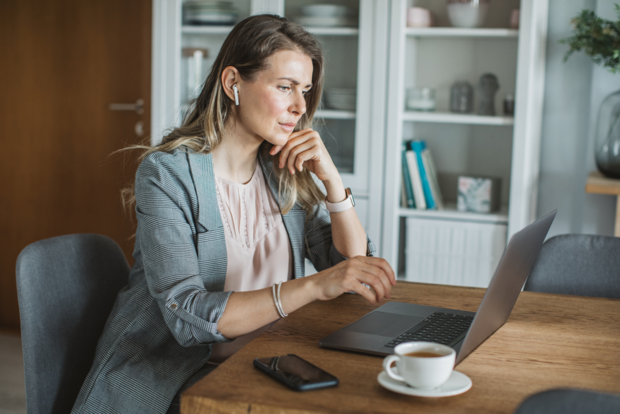 Woman sitting at a table in her home, looking at her open laptop, with a cell phone and coffee next to her