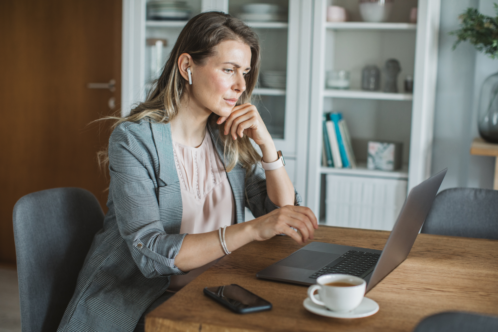 Woman sitting at a table in her home, looking at her open laptop, with a cell phone and coffee next to her