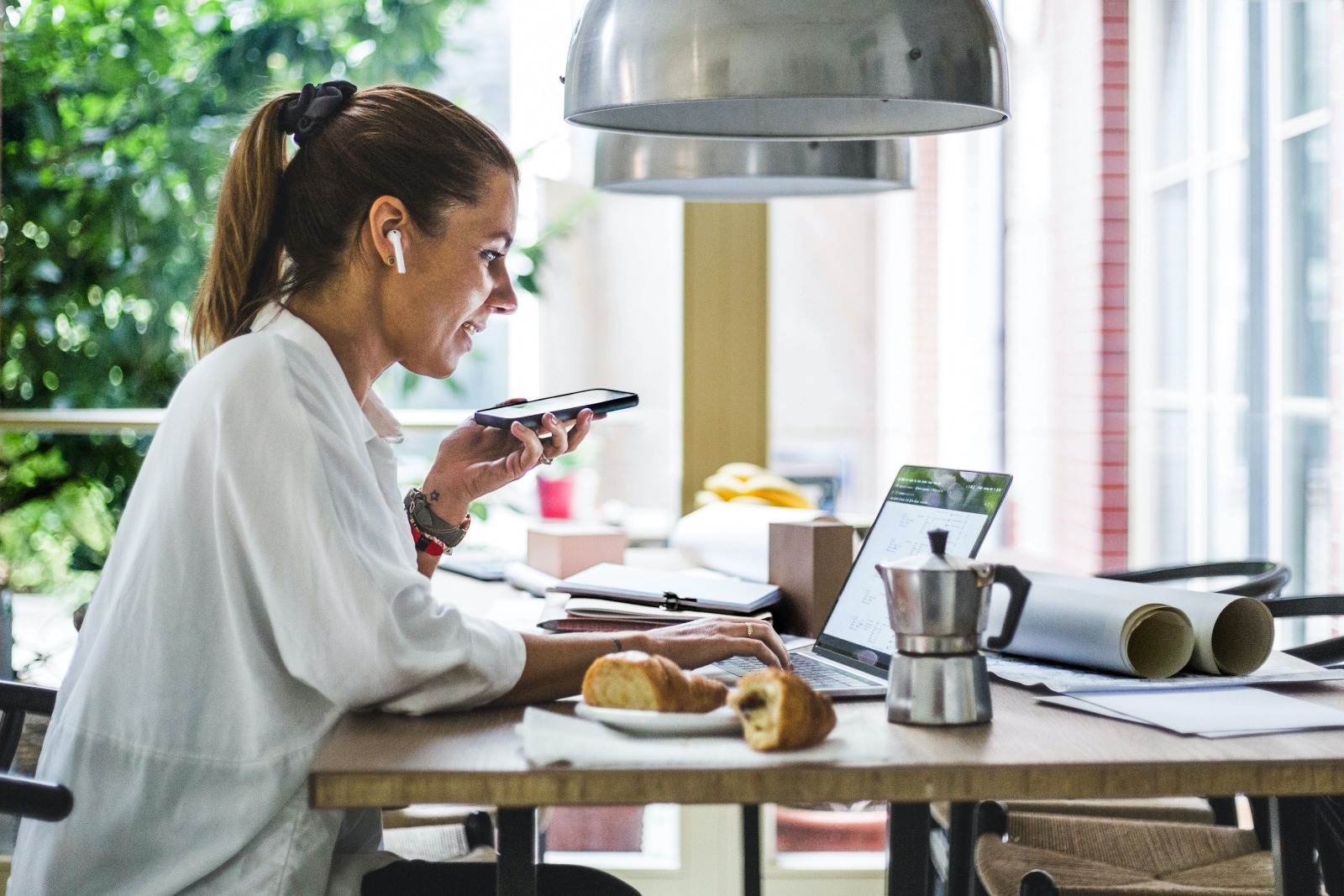 female real estate agent with brown hair in a ponytail and a white flowy top working in her dining room,on her laptop and talking into her cellphone