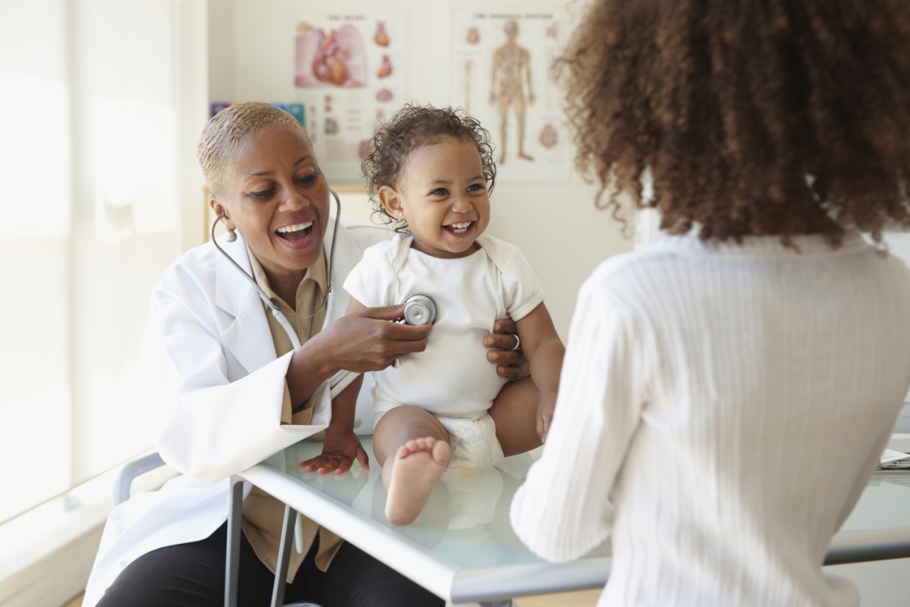 A smiling female doctor uses a stethoscope on a grinning baby who is sitting on an examination table as the baby's mother stands near