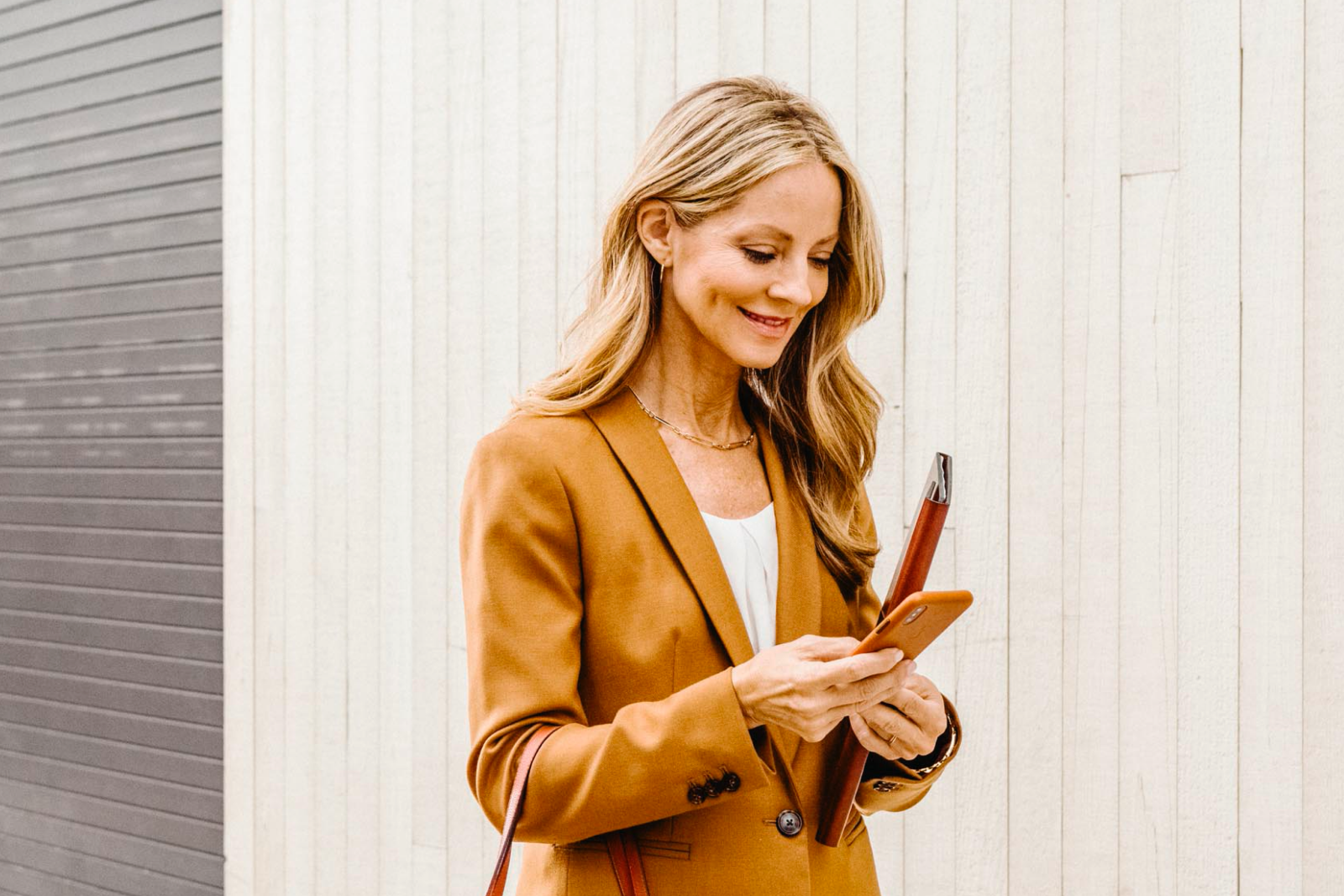 Blonde woman in a mustard blazer looking down at her cell phone, smiling